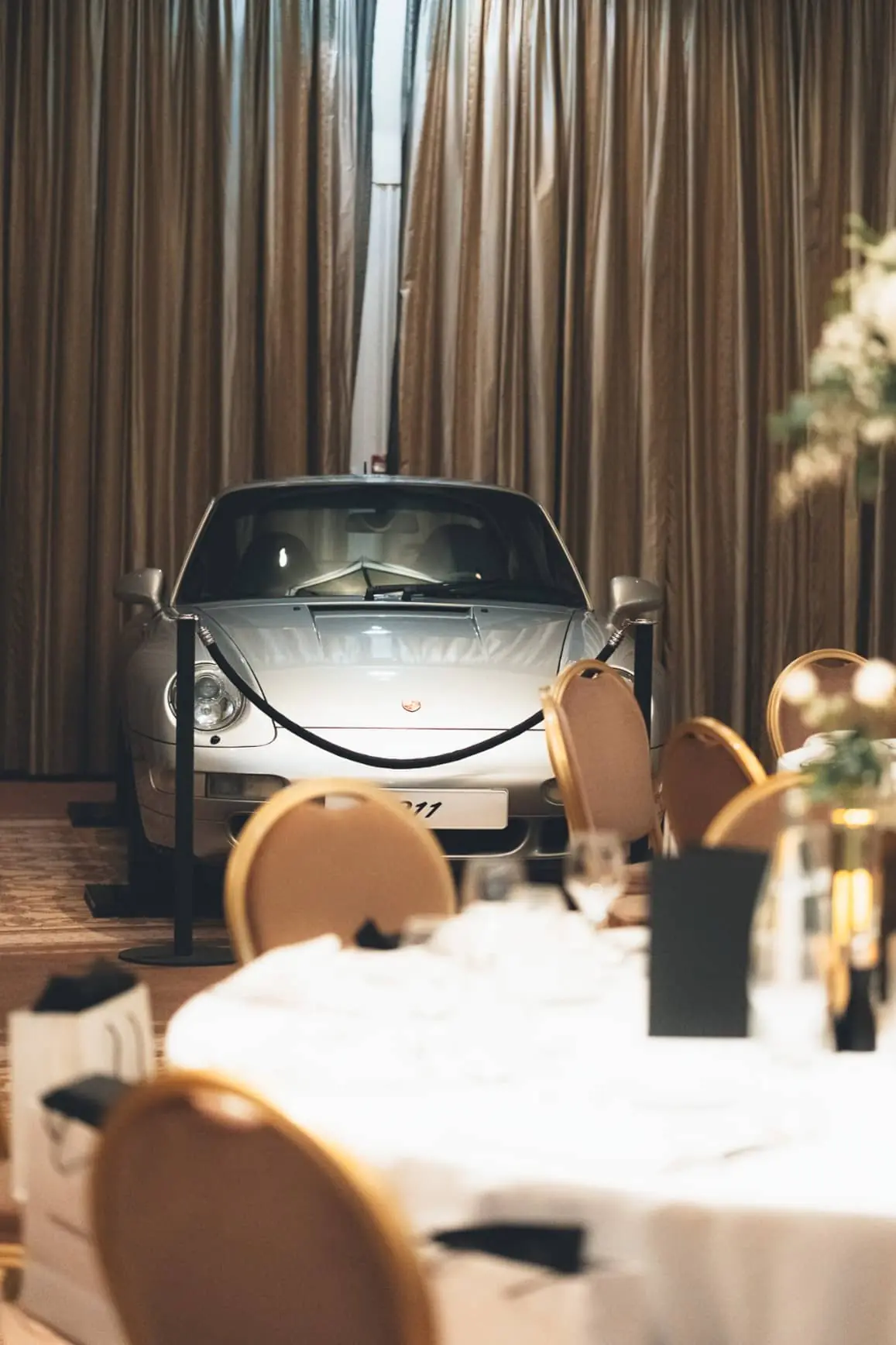 Car displayed in a showroom with thick beige curtains, beige carpet and round tables.