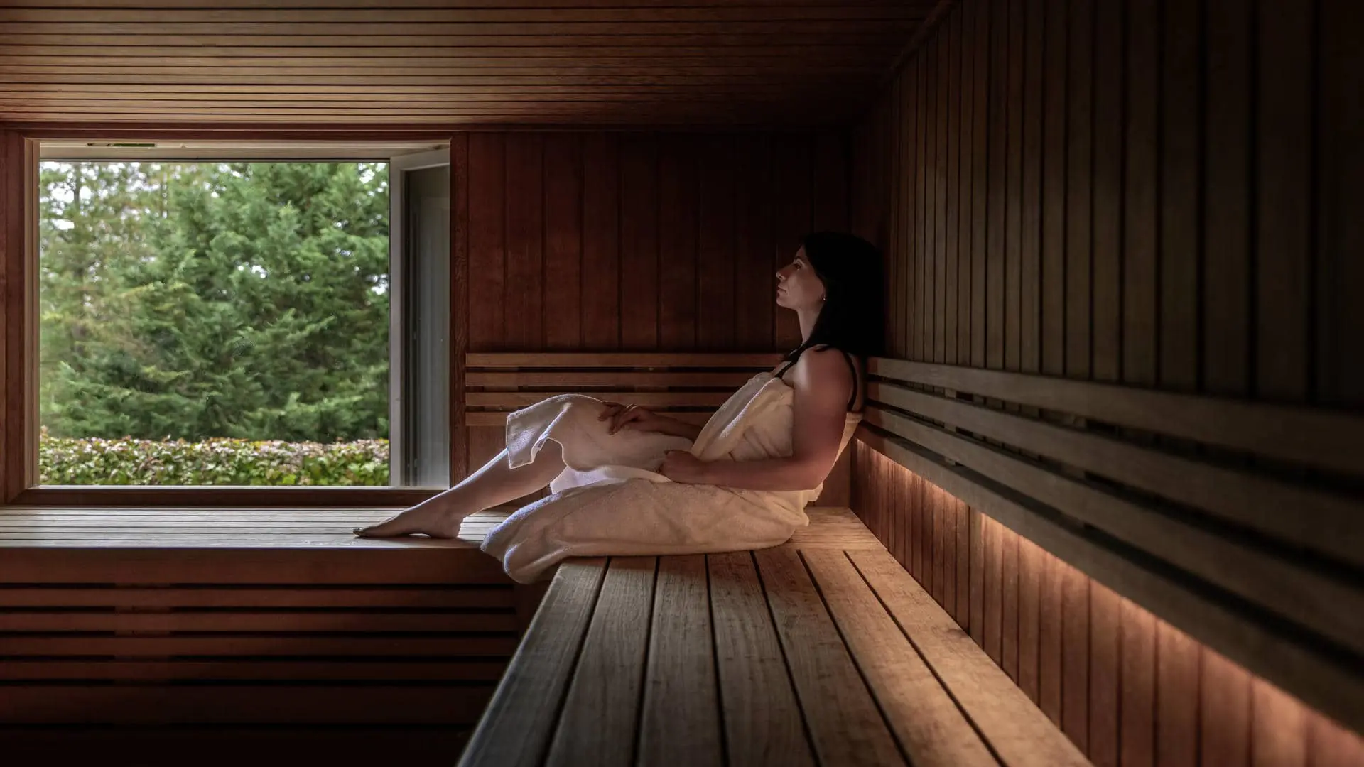 Large all-wood sauna with a view of pine trees and flowering shrubs, featuring a person wrapped in a large white towel.