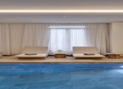 Piscine en carrelage à l’eau bleue, faisant face à de très larges transats avec matelas beiges sur structure en bois, avec en arrière-plan de longs voilages blancs.