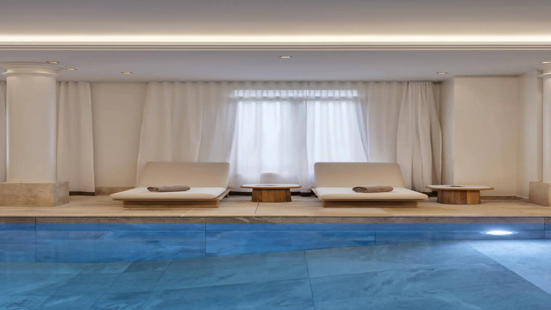 Tiled swimming pool with blue water, facing very wide sun loungers with beige cushions on wooden frames, with full-length white drapes in the background.