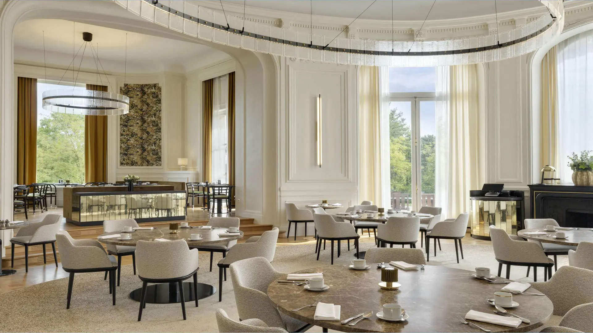 Restaurant room in the non-elevated curved section, with white walls and sheers and curtains opening onto the pine and beech trees and the terrace, large round tables, beige fabric chairs and a designer chandelier on the ceiling.