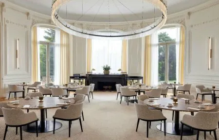 Restaurant room in the non-elevated curved section, with white walls and sheers and curtains opening onto the pine and beech trees and the terrace, large round tables, beige fabric chairs and a designer chandelier on the ceiling.