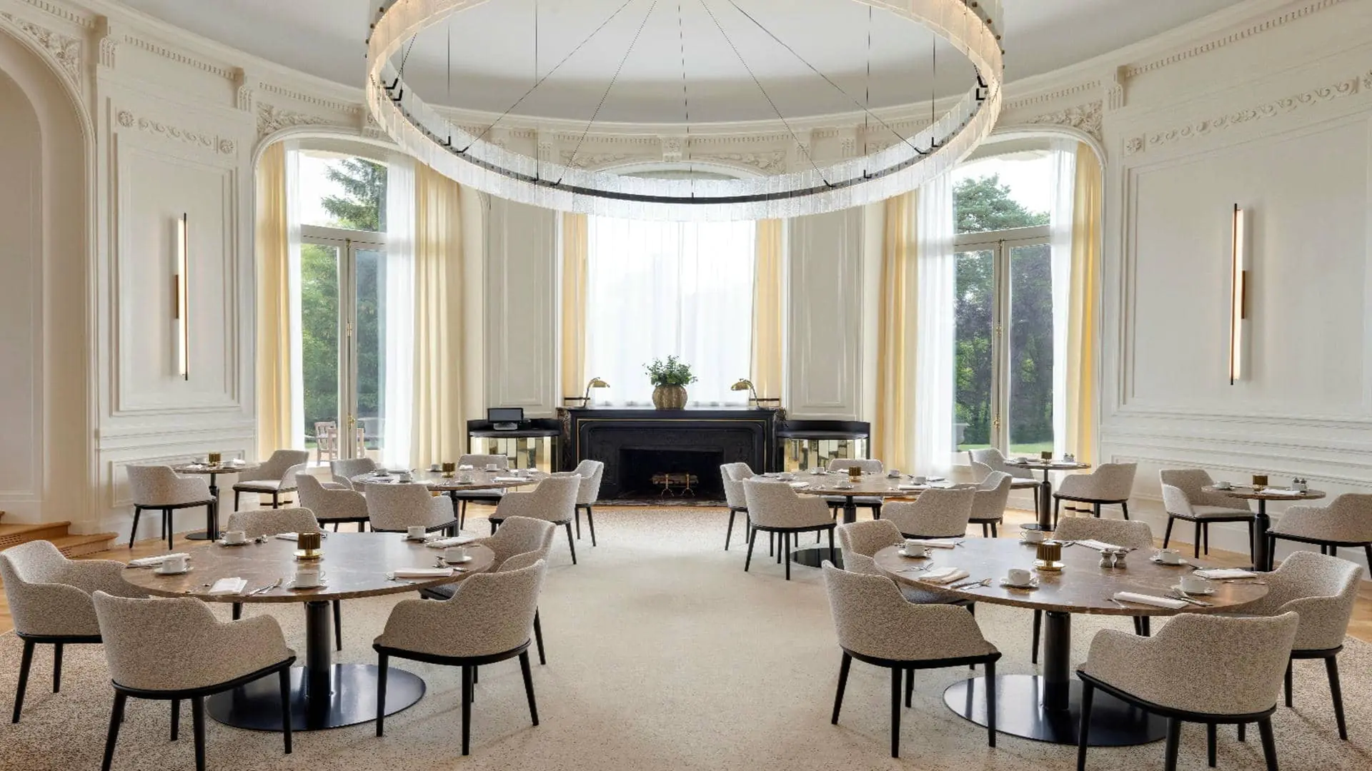 Restaurant room in the non-elevated curved section, with white walls and sheers and curtains opening onto the pine and beech trees and the terrace, large round tables, beige fabric chairs and a designer chandelier on the ceiling.