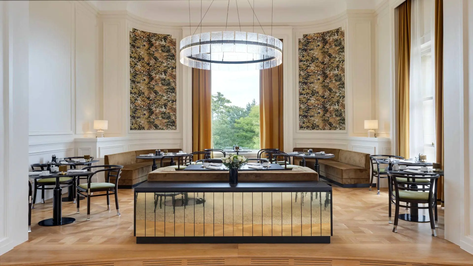 Raised restaurant room with a curved layout, white walls and fabric-trimmed surrounds framing the sheers and curtains opening onto pine and beech trees, brown sofas and black and green chairs around black round tables, with a designer chandelier on the ceiling.