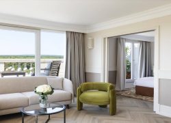 Suite avec vue sur la forêt, salon avec canapé et fauteuils au premier plan, terrasse avec mobilier en arrière-plan, et chambre avec grand tapis sur la droite.