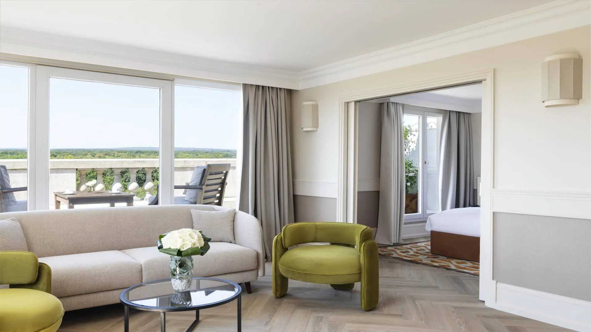 Suite with forest view, featuring a lounge area with sofa and armchairs in the foreground, a terrace with outdoor furniture in the background, and the bedroom with a large rug on the right.