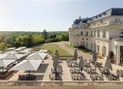 Terrasse extérieure sur dalles de carrelage, avec tables en bois et parasols blancs rectangulaires, ouverts ou fermés, face à la forêt de Chantilly, avec le château sur la gauche.