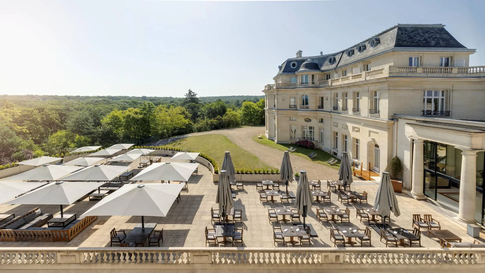 Outdoor terrace with tiled flooring, wooden tables and rectangular white parasols, open or closed, facing the Chantilly forest with the château on the left.
