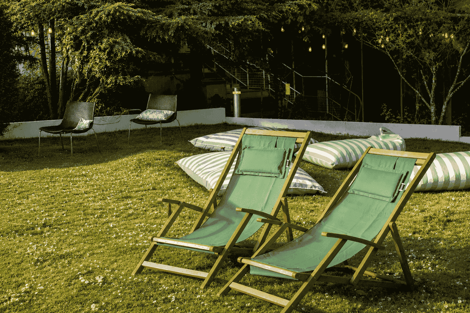 Outdoor lawn area for a countryside seminar with two green wooden deckchairs on the grass in the foreground, striped floor cushions scattered nearby, two woven armchairs beside a low white wall in the background, and trees strung with warm fairy lights above a metal staircase under an evening sky.
