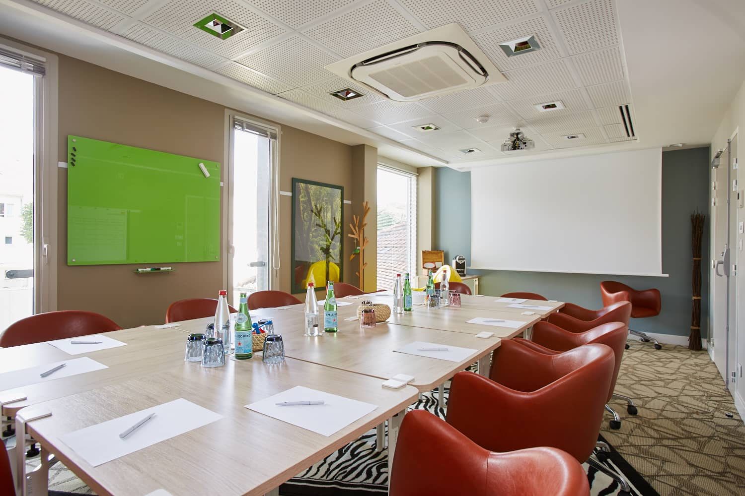 Meeting room for a hôtel pour séminaire with a long light wood conference table set with water bottles, glasses and notepads in the foreground, red leather swivel chairs on both sides, a green glass whiteboard on the left wall, and a large white projection screen against a blue-grey wall in the background.