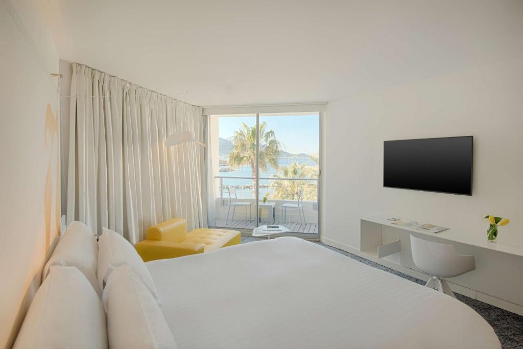 Chambre d'hôtel pour séminaire en bord de mer, avec un grand lit blanc au premier plan, une méridienne jaune à côté d'un lampadaire à gauche, une télévision murale et un bureau blanc à droite, et un balcon donnant sur des palmiers et la mer en arrière-plan.