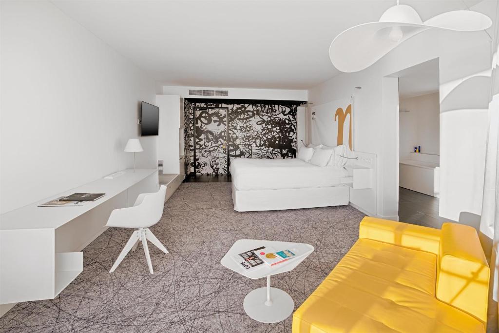 Chambre d'hôtel pour hébergement groupe en vue générale avec un lit blanc à l'arrière-plan, un long bureau blanc et une chaise à gauche, un canapé jaune et une table basse blanche au premier plan à droite, une moquette à motif graphique, et un mur décoratif noir et blanc derrière le lit.