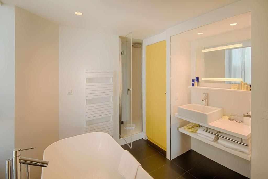 Salle de bain d'hôtel pour hébergement groupe, avec une baignoire îlot blanche au premier plan à gauche, une cabine de douche vitrée et un sèche-serviettes blanc au centre, un meuble vasque suspendu avec lavabo carré et miroir lumineux à droite, et un panneau mural jaune au-dessus d'un sol carrelé foncé en arrière-plan.