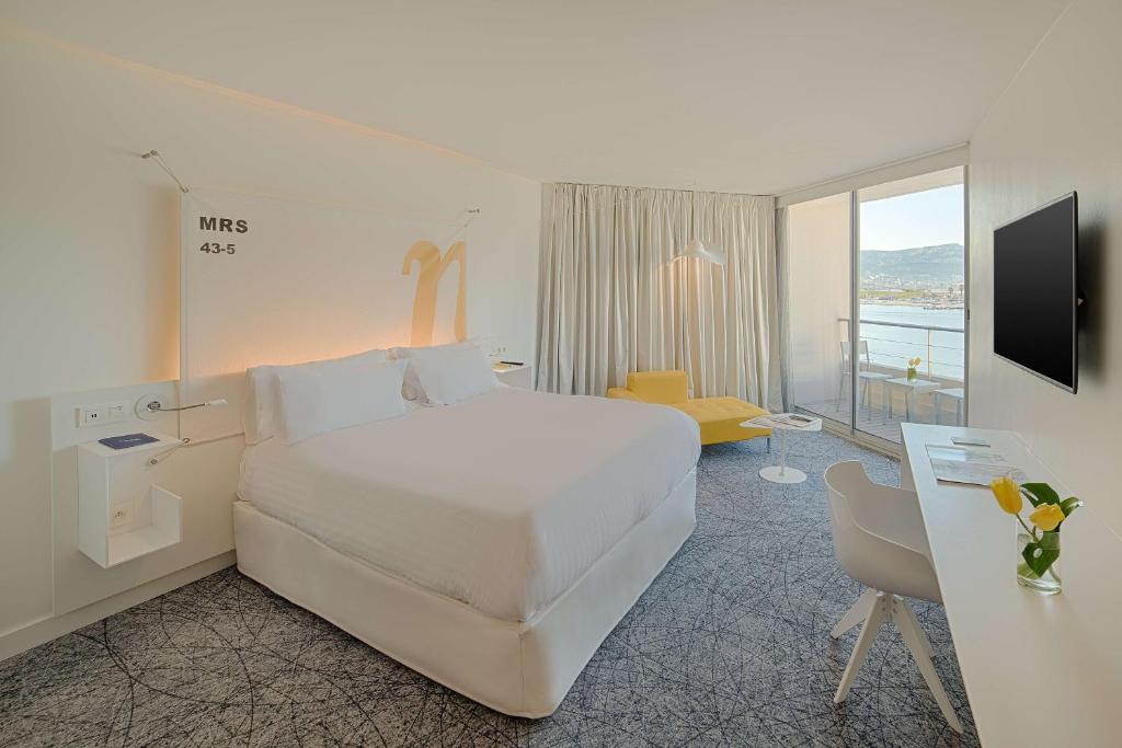 Chambre d'hôtel pour séminaire en bord de mer, avec un grand lit blanc à gauche au premier plan, un bureau blanc avec chaise à droite, un fauteuil jaune avec une petite table ronde au centre, et un balcon avec vue sur la mer et les montagnes en arrière-plan.
