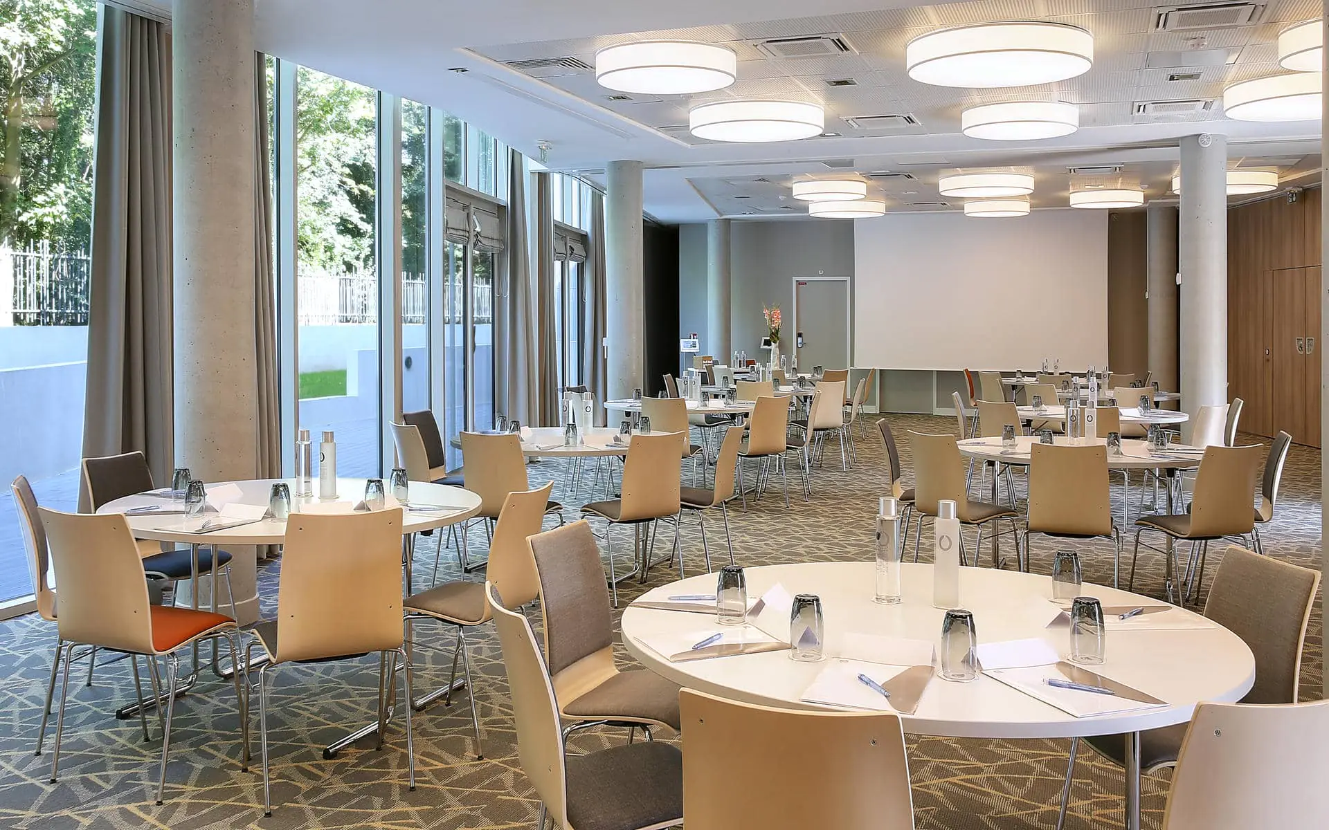 Seminar room for location de salle with round white tables set with water bottles, glasses and notepads surrounded by beige and grey upholstered chairs in the foreground, a large white projection screen against a beige wall in the background, round drum ceiling lights above, and floor-to-ceiling windows on the left revealing trees outside.