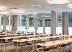 Salle de formation avec rangées de tables blanches rectangulaires dressées de bouteilles d'eau, verres et blocs-notes au premier plan, des chaises capitonnées foncées de part et d'autre, des suspensions rondes en forme de tambour au plafond, et de grandes baies vitrées à rideaux beiges laissant apercevoir un jardin clôturé et des arbres à l'arrière-plan.