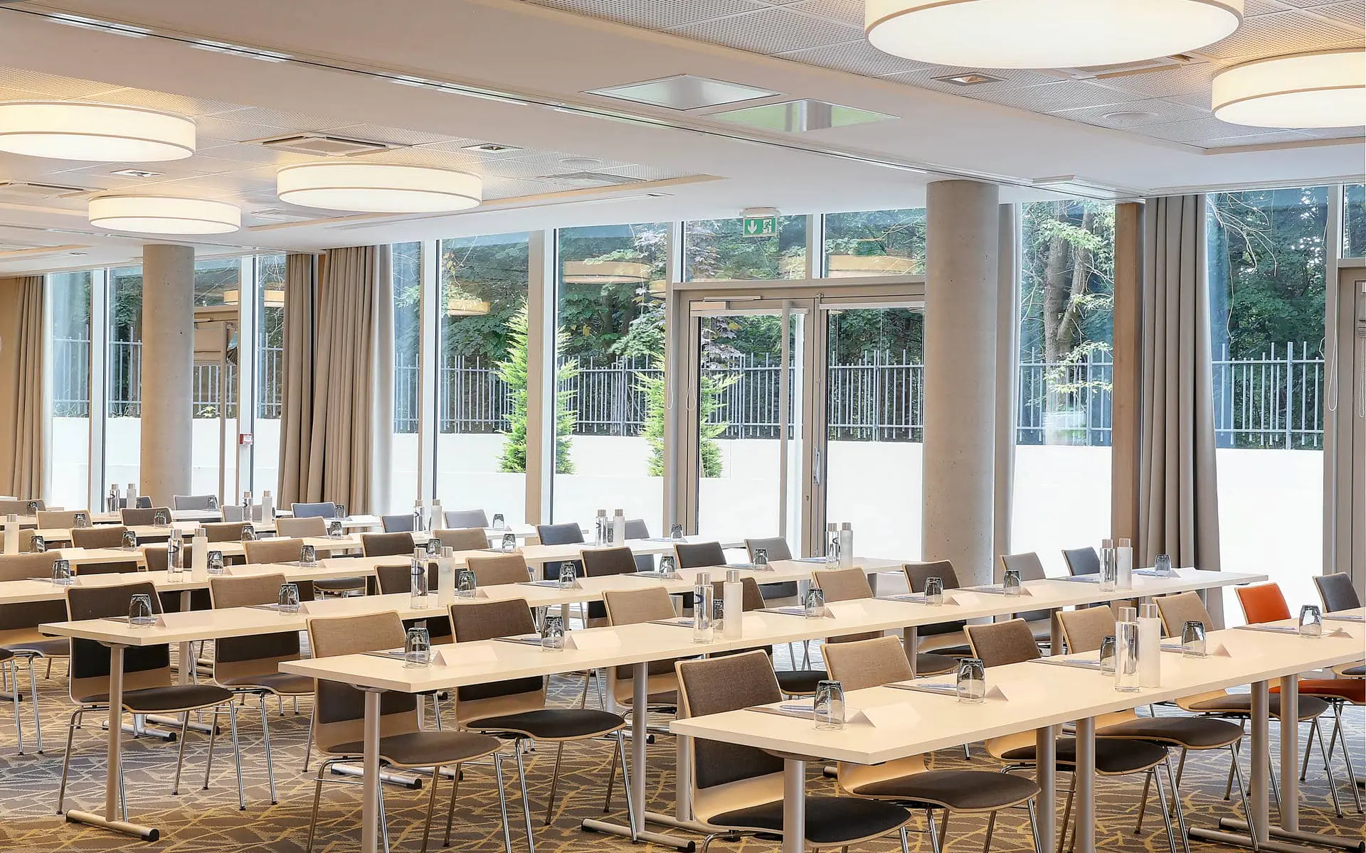 Training room with rows of white rectangular tables set with water bottles, glasses and notepads in the foreground, dark upholstered chairs on either side, round drum ceiling lights above, and floor-to-ceiling windows with beige curtains revealing a fenced garden and trees in the background.