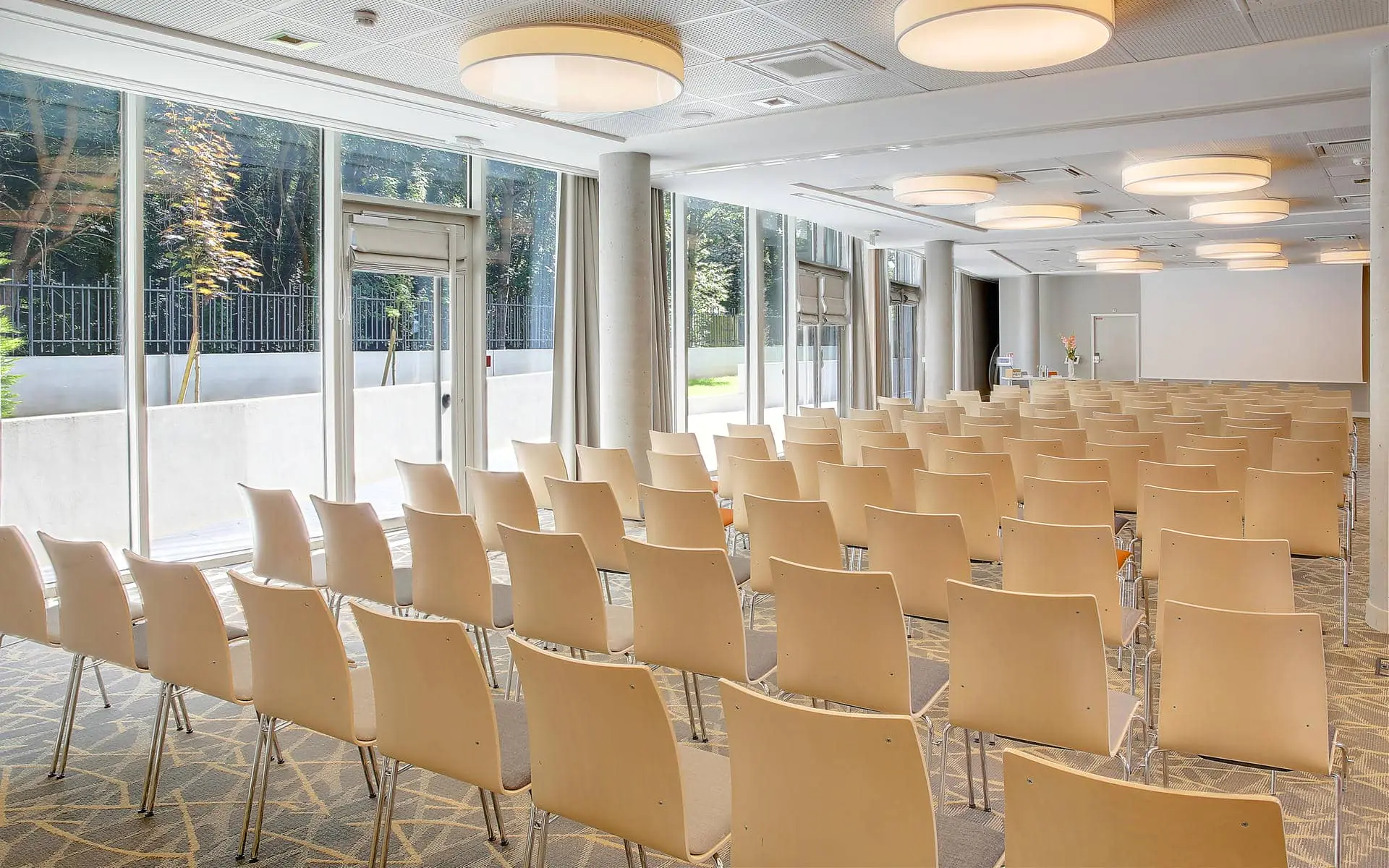 Large training room set up for a hôtel pour séminaire with rows of light wood and beige chairs facing forward in the foreground, round drum pendant lights hanging from a white ceiling, stone columns dividing the space, and floor-to-ceiling windows on the left revealing a garden with trees under a stone perimeter wall in the background.