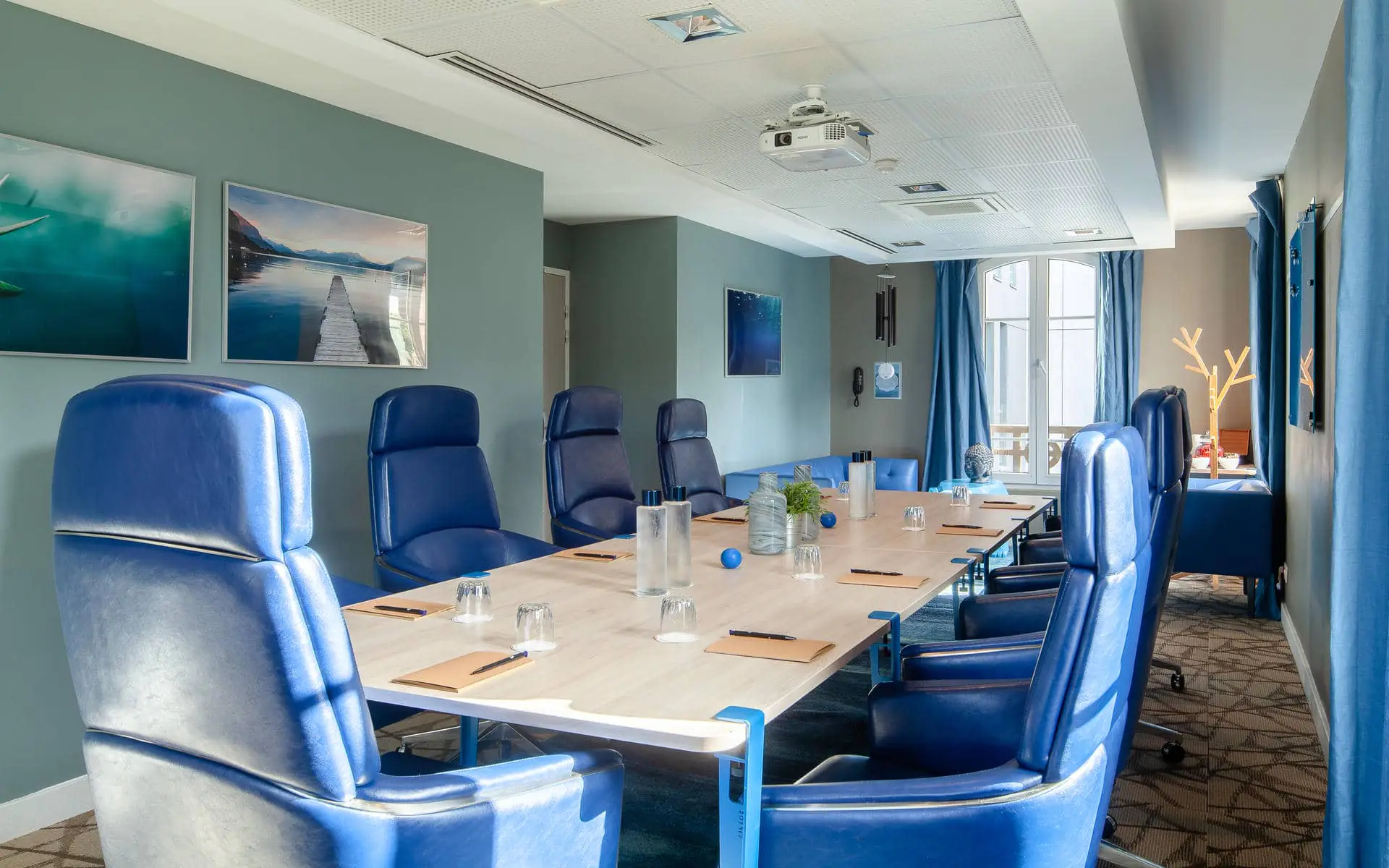 Meeting room for training rooms with a long light wood conference table set with notepads, pens and water glasses in the foreground, high-backed blue leather swivel chairs on both sides, framed lake landscape photographs on a sage green wall to the left, and blue curtained windows with a lounge seating area visible in the background.
