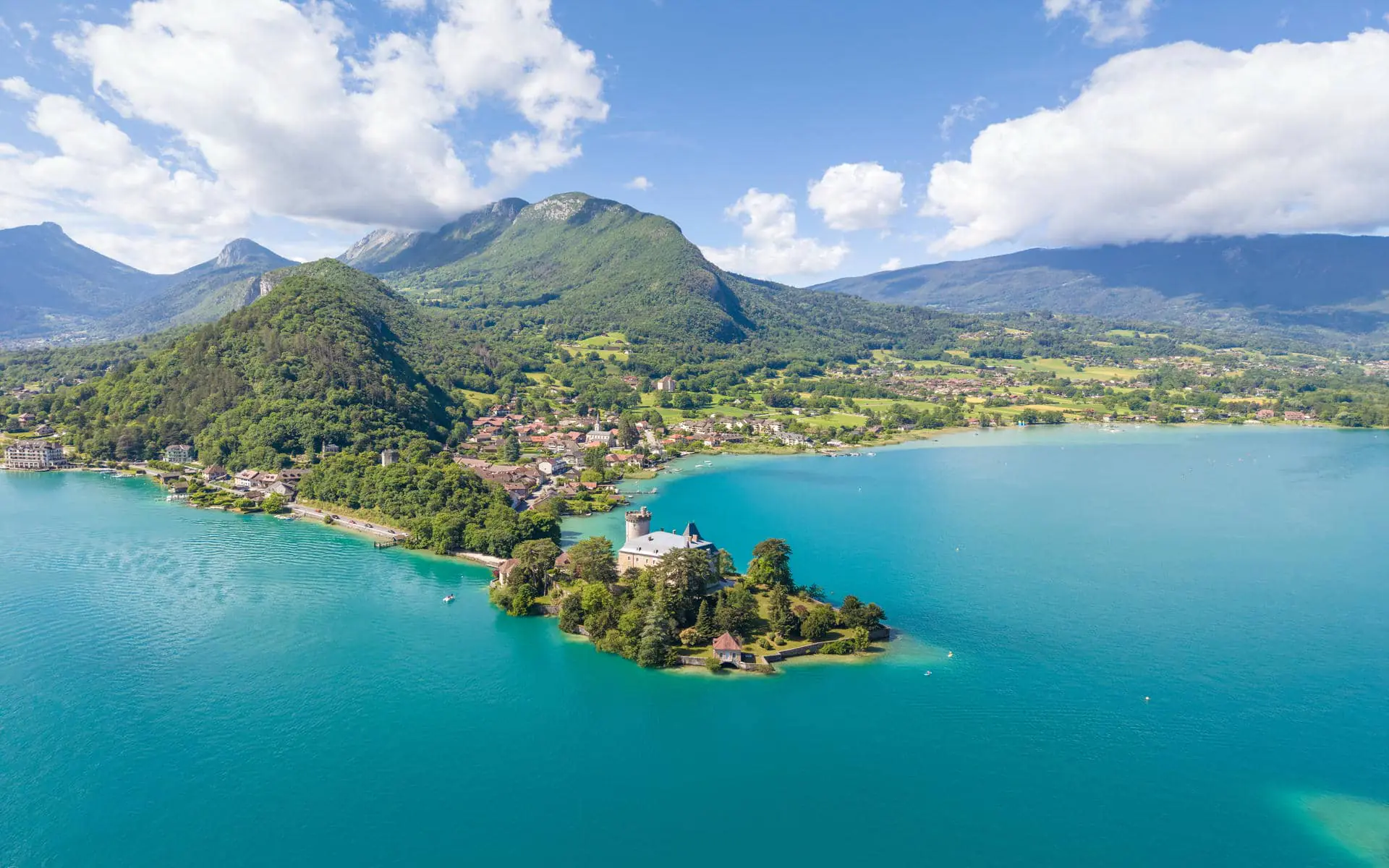 Vue aérienne du lac d'Annecy avec un lac turquoise entourant une presqu'île boisée où se dresse un château en pierre à tour ronde au premier plan, village lacustre aux maisons à toits rouges et clocher d'église à gauche, et montagnes boisées verdoyantes s'élevant sous un ciel partiellement nuageux en arrière-plan, un paysage adapté à un séminaire au vert.