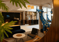 Hall d'hôtel pour hébergement de groupe avec fauteuils en velours bleu marine et moutarde autour d'une table basse ronde en marbre au premier plan, des statues décoratives de girafes fleuries bleues placées près d'un mur en bois à droite, une banque d'accueil avec étagères ouvertes en bois à l'arrière-plan, et des feuilles de palmier vertes encadrant la prise de vue au premier plan à gauche.