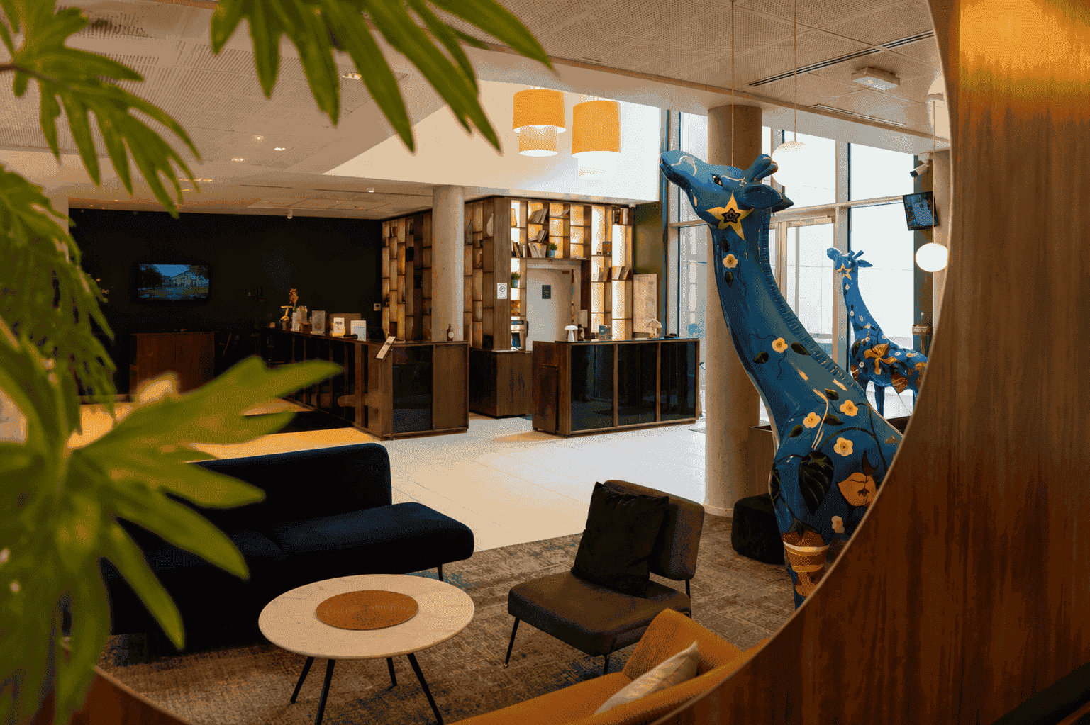 Hotel lobby for group accommodation with dark blue and mustard velvet armchairs around a round marble coffee table in the foreground, decorative blue floral giraffe statues standing beside a wood-panelled wall on the right, a reception desk with open wooden shelving in the background, and green palm leaves framing the shot in the foreground on the left.
