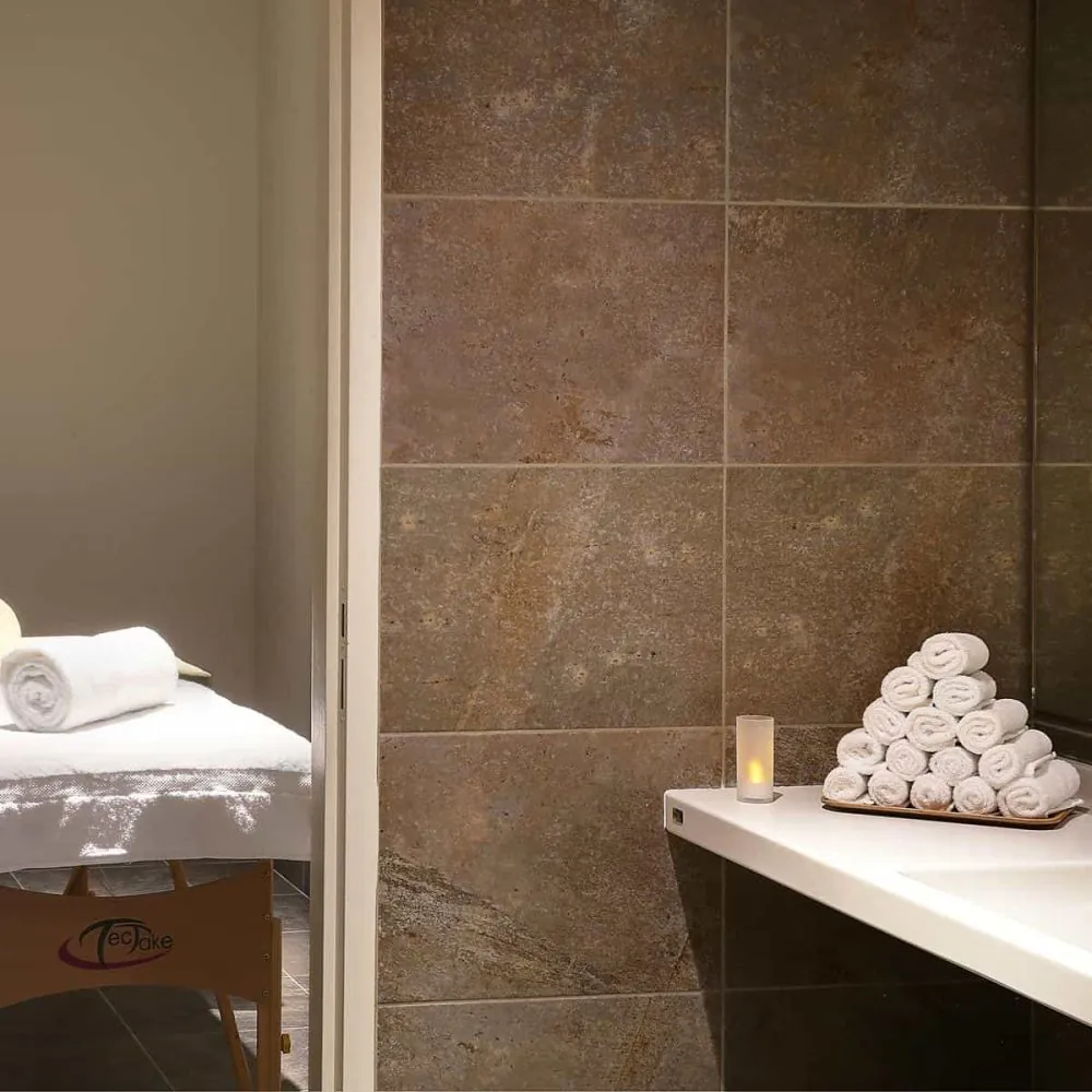 Wellness seminar space with a massage table dressed in white linen and a rolled towel on the left, an amber candle and orchid on a small side table nearby, a stacked pyramid of rolled white towels on a marble sink counter beside soap dispensers on the right, and dark textured stone-tiled walls under warm lighting.