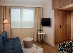 en:: lounge area of a seminar hotel suite in Lyon, with a blue sofa, beige curtains, wooden wardrobe, black-and-white armchair, and a small white table with flowers fr:: espace salon d’une suite d’hôtel séminaire à Lyon, avec canapé bleu, rideaux beiges, armoire en bois, fauteuil noir et blanc et petite table blanche fleurie