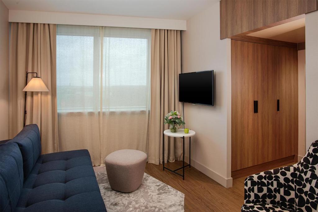 en:: lounge area of a seminar hotel suite in Lyon, with a blue sofa, beige curtains, wooden wardrobe, black-and-white armchair, and a small white table with flowers fr:: espace salon d’une suite d’hôtel séminaire à Lyon, avec canapé bleu, rideaux beiges, armoire en bois, fauteuil noir et blanc et petite table blanche fleurie
