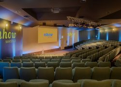 Auditorium pensé pour un séminaire en bord de mer de grande envergure, comprenant des rangées de sièges gradins vert pâle sur un sol sombre à motifs au premier plan, une scène avec pupitre et écran de projection affichant le logo « nhow Marseille » au centre, et des murs éclairés en bleu avec la marque « nhow Marseille » à gauche.