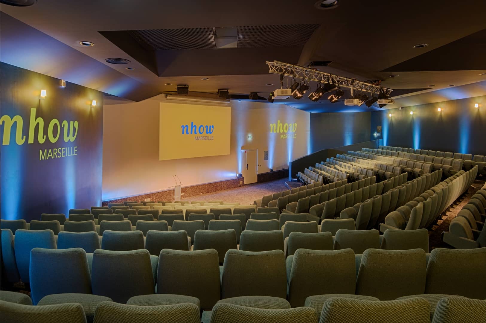 Auditorium pensé pour un séminaire en bord de mer de grande envergure, comprenant des rangées de sièges gradins vert pâle sur un sol sombre à motifs au premier plan, une scène avec pupitre et écran de projection affichant le logo « nhow Marseille » au centre, et des murs éclairés en bleu avec la marque « nhow Marseille » à gauche.