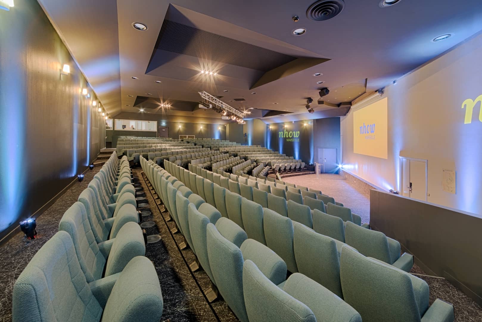 Auditorium pensé pour un séminaire en ville de grande envergure, comprenant des rangées de sièges gradins vert pâle sur un sol sombre à motifs au premier plan, une scène avec écran de projection affichant le logo « nhow Marseille » au centre, et des murs éclairés en bleu avec projecteurs en arrière-plan.