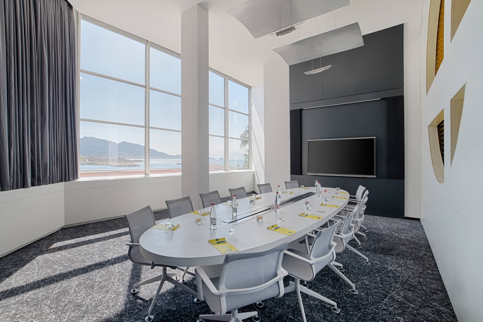 Salle de conseil pensée pour un séminaire en bord de mer, comprenant une table ovale blanche avec fauteuils pivotants gris, bouteilles d'eau et pochettes sur une moquette sombre à motifs au premier plan, un grand écran intégré à un panneau mural gris foncé au centre, et de grandes baies vitrées donnant sur la mer et les montagnes à gauche.