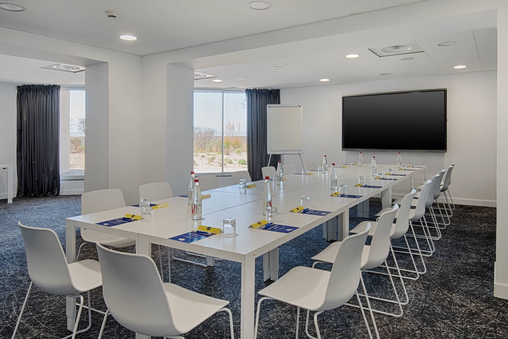 Salle de réunion pensée pour un séminaire en ville, comprenant une longue table blanche avec chaises blanches, bouteilles d'eau et pochettes sur une moquette sombre à motifs au premier plan, un écran mural et un paperboard au centre, et des fenêtres aux rideaux foncés donnant sur un paysage naturel à gauche.