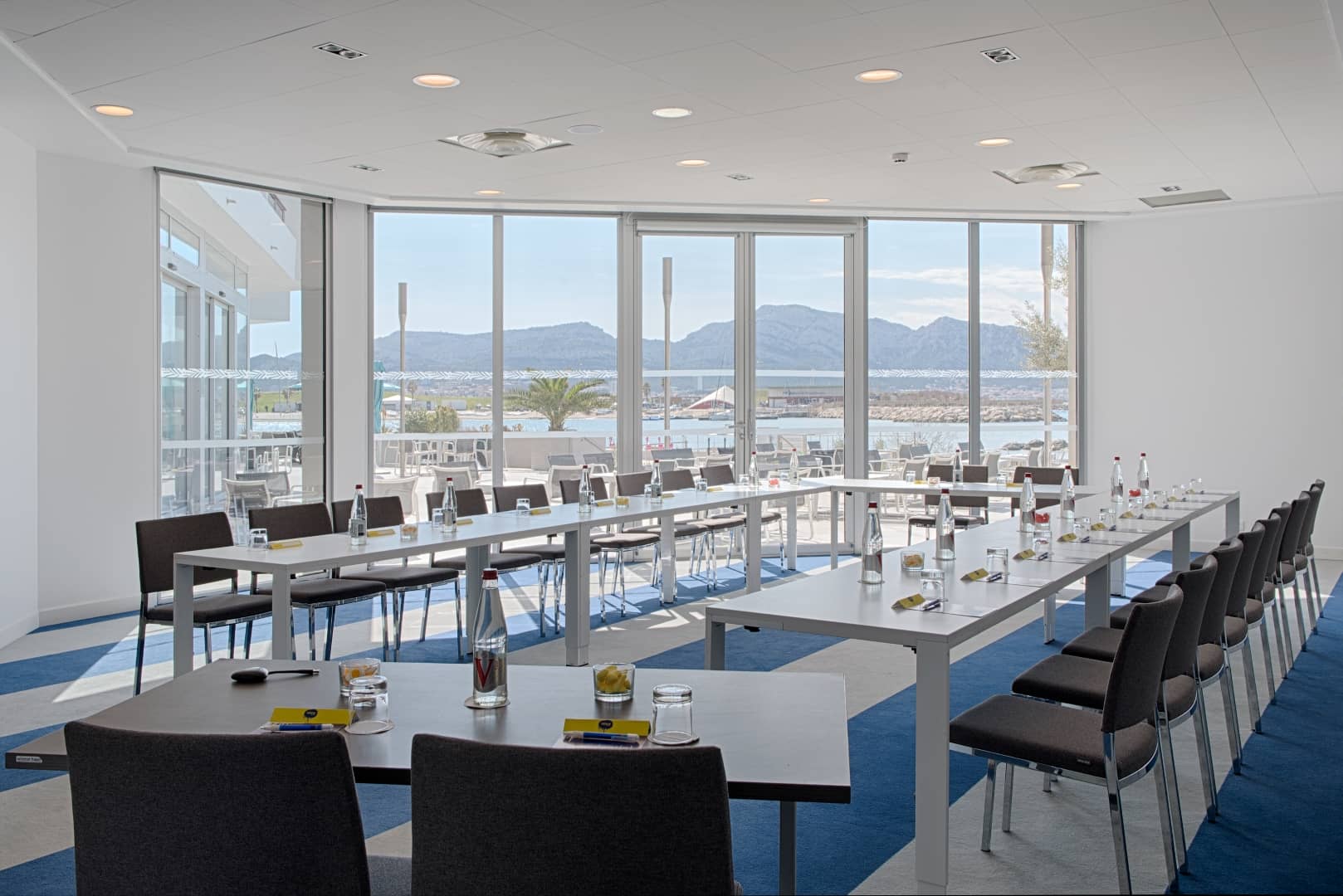 Salle de réunion installée en rangées pour un séminaire en ville, comprenant des tables blanches avec chaises marron, bouteilles d'eau et verres sur une moquette rayée bleu marine et beige au premier plan, et de grandes baies vitrées donnant sur un littoral montagneux avec marina en arrière-plan.