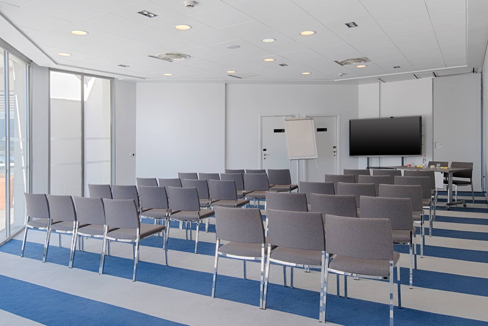 Salle de conférence installée en configuration théâtre pour un séminaire en ville, comprenant des rangées de chaises grises sur une moquette rayée bleu marine et beige au premier plan, un écran mural et un paperboard au centre, et de grandes baies vitrées à gauche.