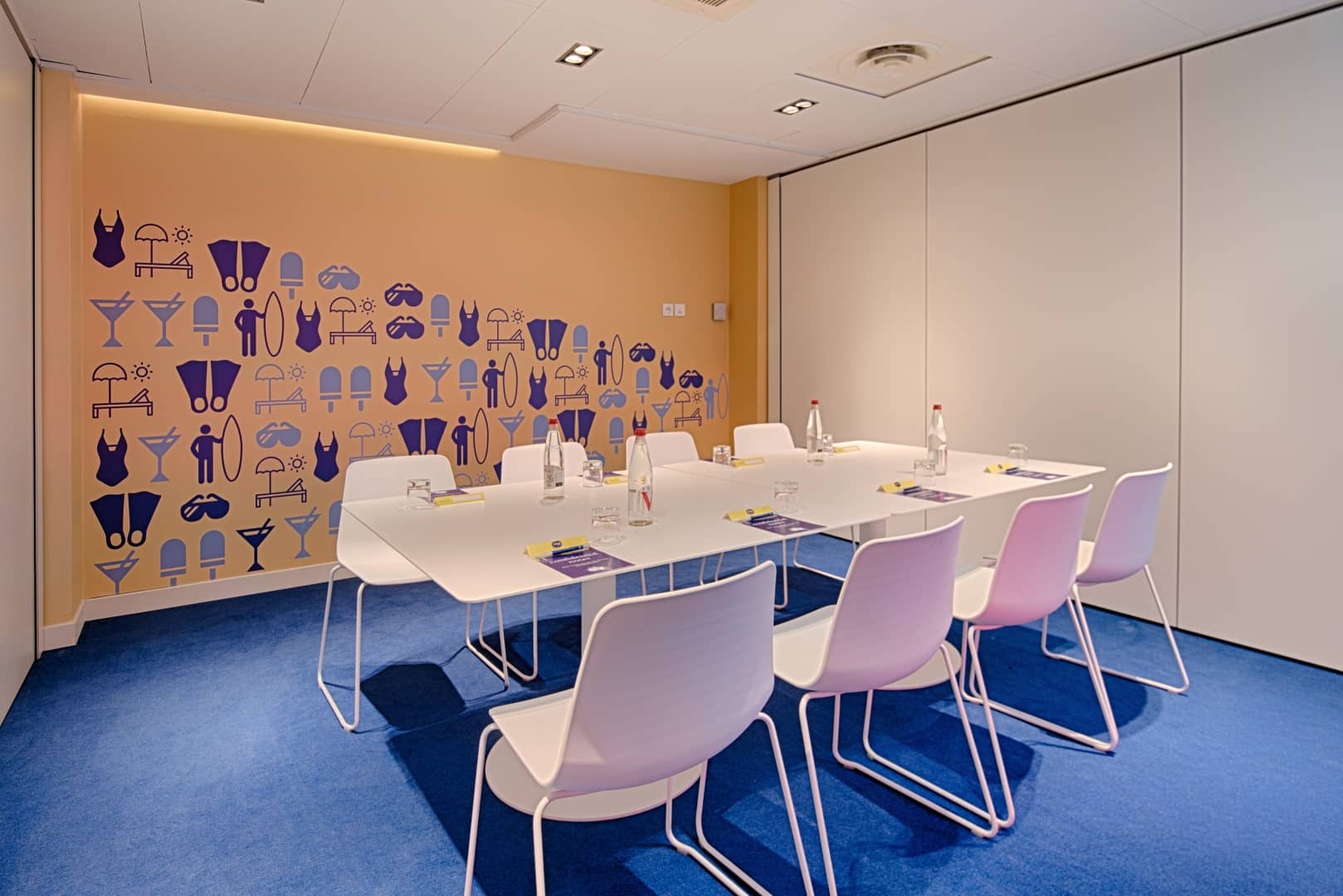 Salle de réunion évoquant une option de location de salle, comprenant une table rectangulaire blanche avec chaises roses pastel et blanches, bouteilles d'eau et pochettes sur une moquette bleue au premier plan, et un mur orange recouvert d'illustrations estivales de maillots de bain, cocktails et tongs en arrière-plan.