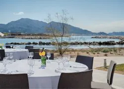 Terrasse de restauration extérieure s'inscrivant dans l'offre de restauration sur place, comprenant des tables rondes nappées de blanc avec verres à vin et tulipes jaunes au premier plan, des chaises gris foncé autour des tables au centre, et un littoral rocheux avec montagnes et baie calme en arrière-plan.