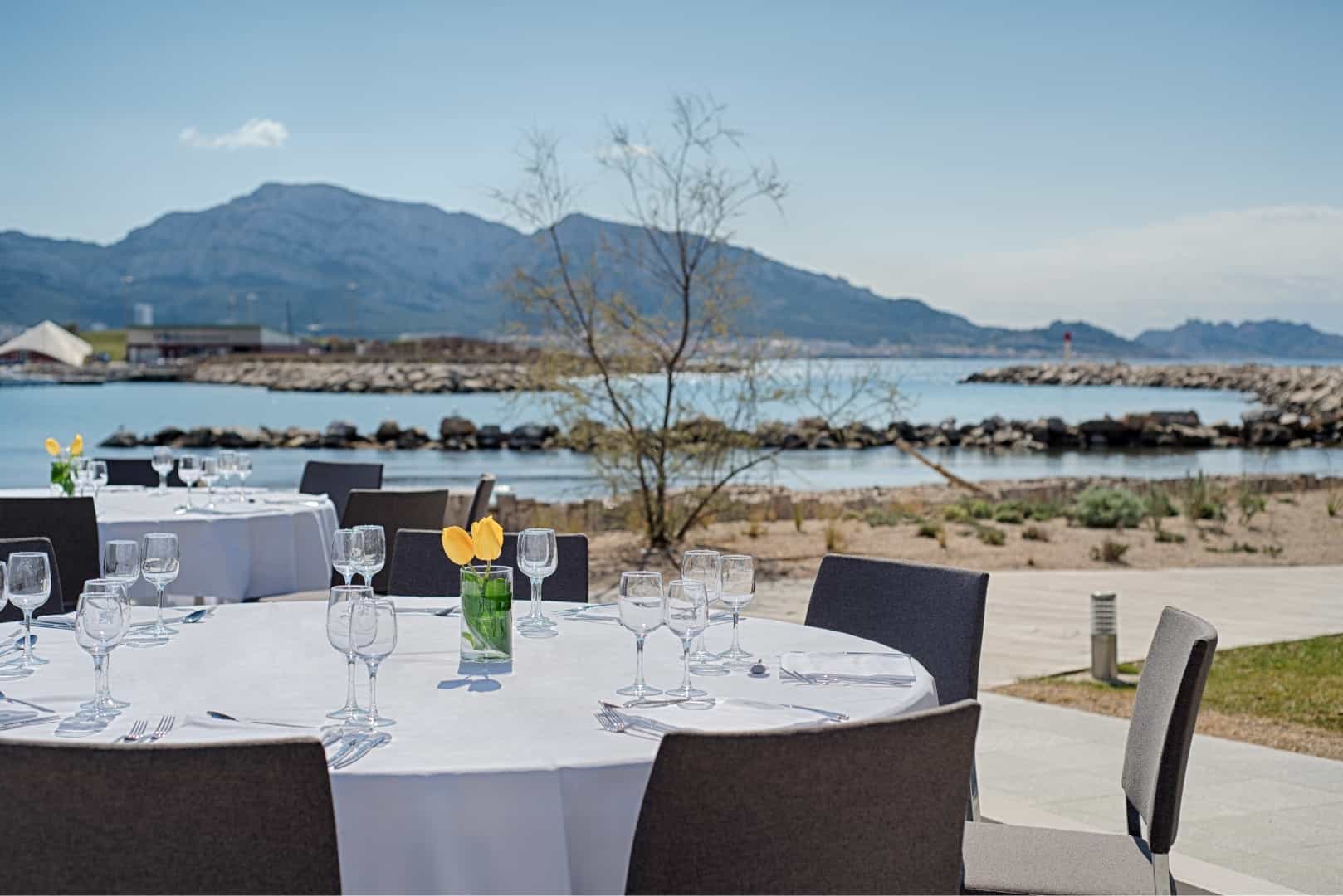 Terrasse de restauration extérieure s'inscrivant dans l'offre de restauration sur place, comprenant des tables rondes nappées de blanc avec verres à vin et tulipes jaunes au premier plan, des chaises gris foncé autour des tables au centre, et un littoral rocheux avec montagnes et baie calme en arrière-plan.