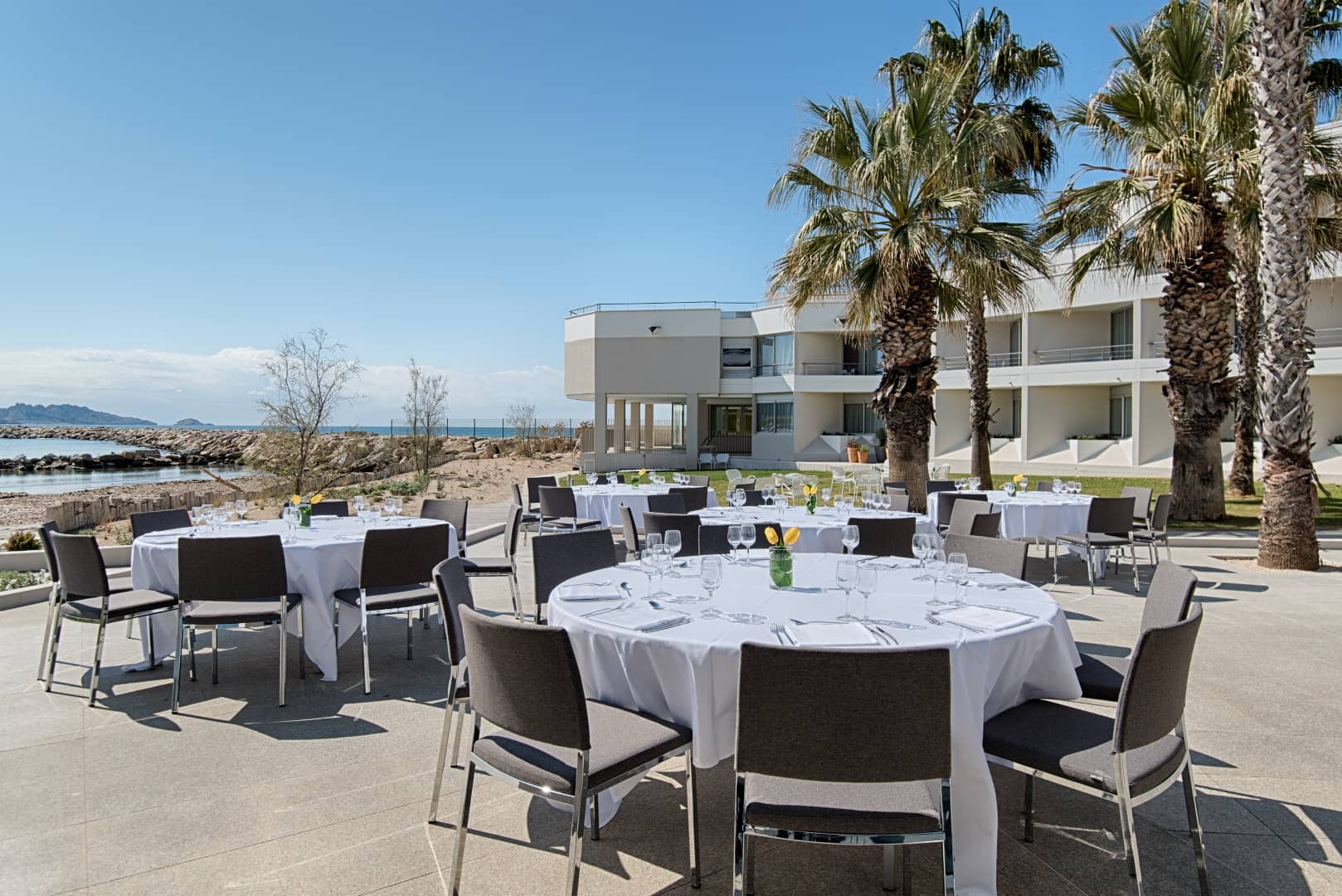Terrasse d'hôtel pour restauration sur place avec des tables rondes recouvertes de nappes blanches entourées de chaises grises au premier plan, des palmiers et un bâtiment d'hôtel contemporain à droite, la mer et un rivage rocheux à l'arrière-plan à gauche, sous un ciel bleu dégagé.