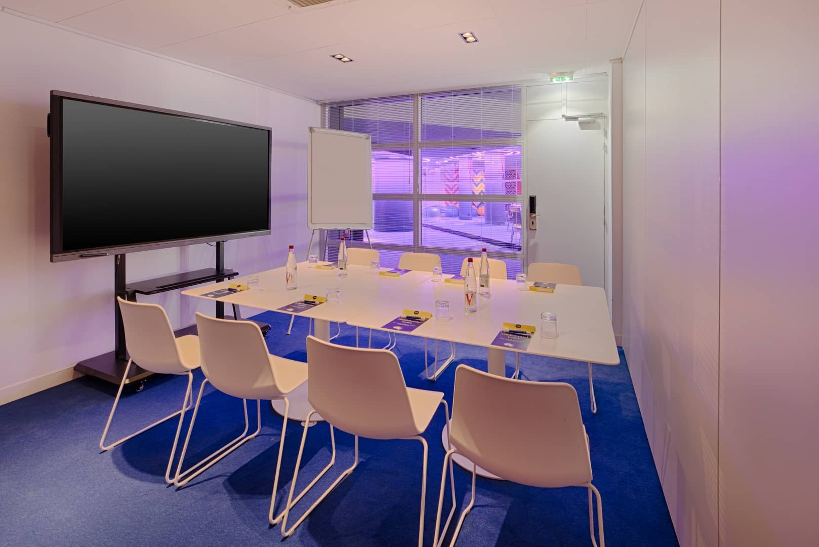 Petite salle de réunion proposée pour une option de location de salle, comprenant une table rectangulaire blanche avec chaises beiges, bouteilles d'eau et pochettes sur une moquette bleue au premier plan, un écran mobile et un paperboard à gauche, et une fenêtre à stores donnant sur un couloir éclairé en violet en arrière-plan.