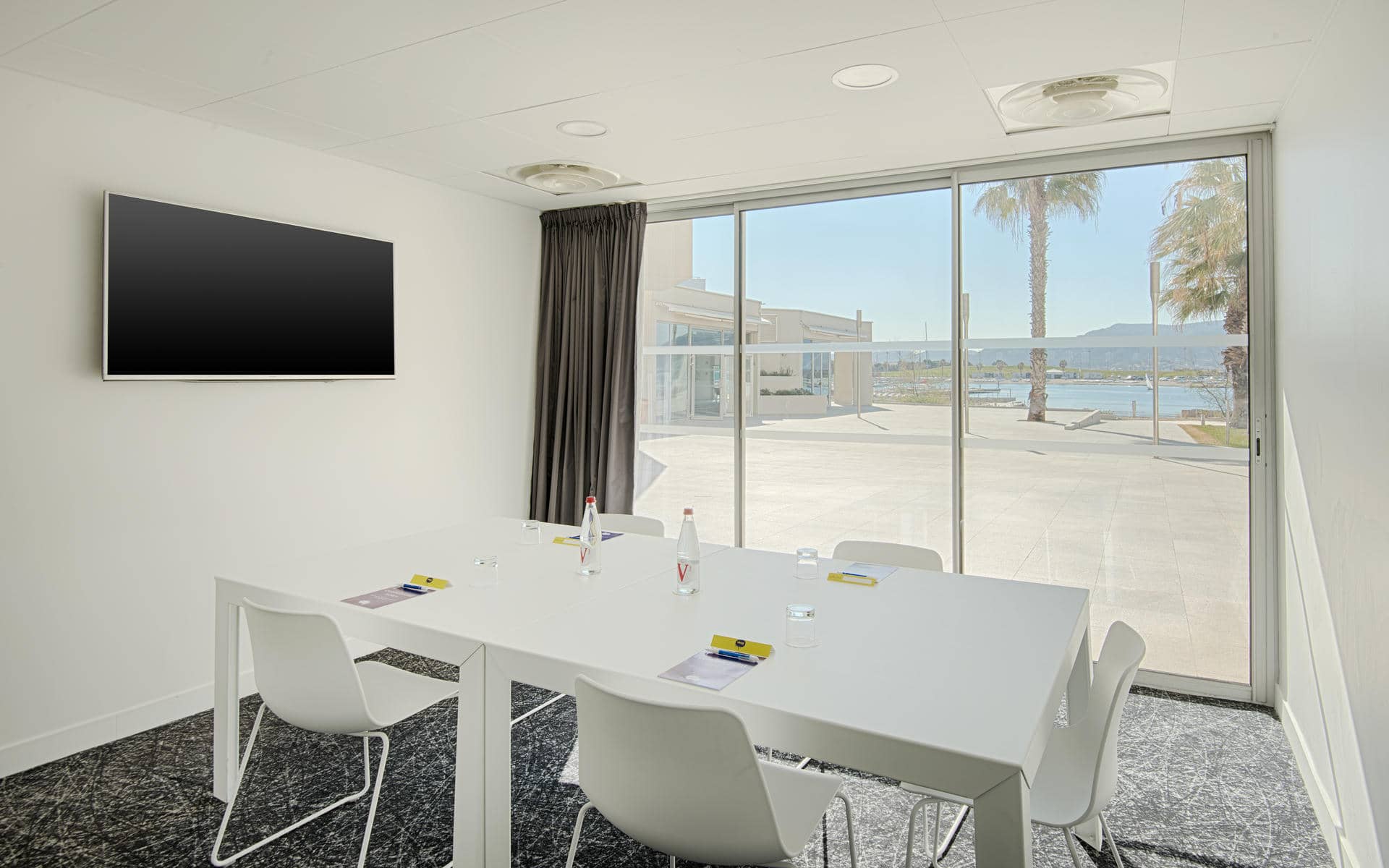 Petite salle de réunion proposant une option de location de salle, comprenant une table rectangulaire blanche avec chaises blanches, bouteilles d'eau et blocs-notes sur une moquette sombre à motifs au premier plan, un écran mural à gauche, et une grande baie vitrée ouvrant sur une terrasse en bord d'eau avec palmiers en arrière-plan.
