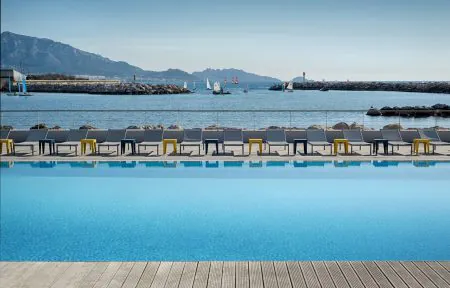 Piscine extérieure d'hôtel adaptée pour un séminaire en bord de mer, comprenant une piscine à débordement bleu vif avec terrasse en bois au premier plan, une rangée de chaises longues grises avec tables d'appoint jaunes et bleues le long d'une rambarde vitrée au centre, et la mer avec voiliers et montagnes sous un ciel dégagé en arrière-plan.