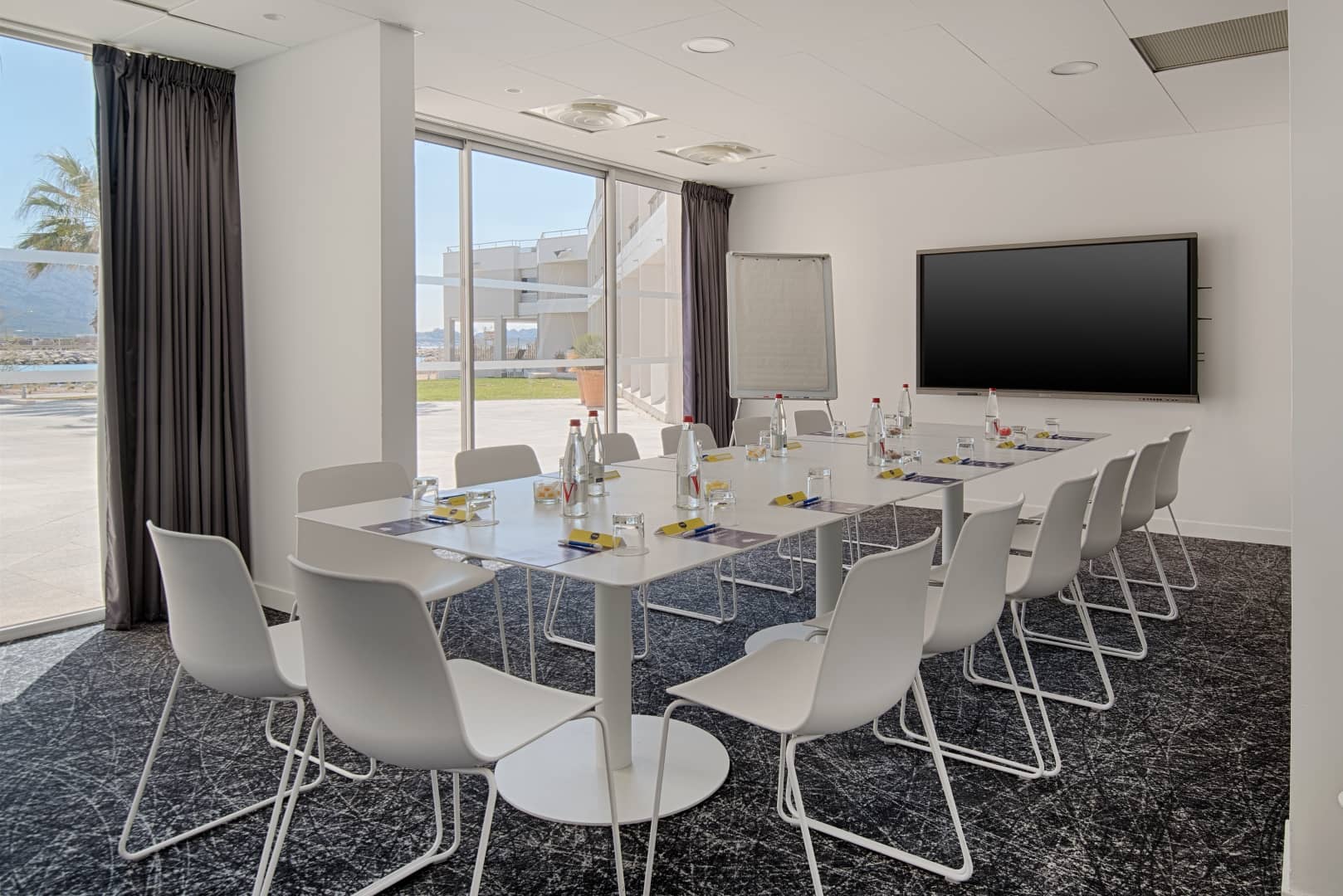 Salle de réunion pensée pour un séminaire en ville, comprenant une table rectangulaire blanche avec chaises blanches, bouteilles d'eau et blocs-notes sur une moquette sombre à motifs au premier plan, un paperboard et un grand écran mural au centre, et de grandes baies vitrées donnant sur une terrasse et le littoral à gauche.