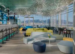 Lounge bar en rooftop propice à un moment afterwork, comprenant des canapés incurvés bleu pâle avec pouf bleu marine et petite table ronde au premier plan, une banquette modulable jaune-vert au centre, un comptoir de bar avec tabourets à gauche, et une nuée de sculptures de poissons suspendues au plafond avec vue sur mer et montagnes à travers de grandes baies vitrées en arrière-plan.