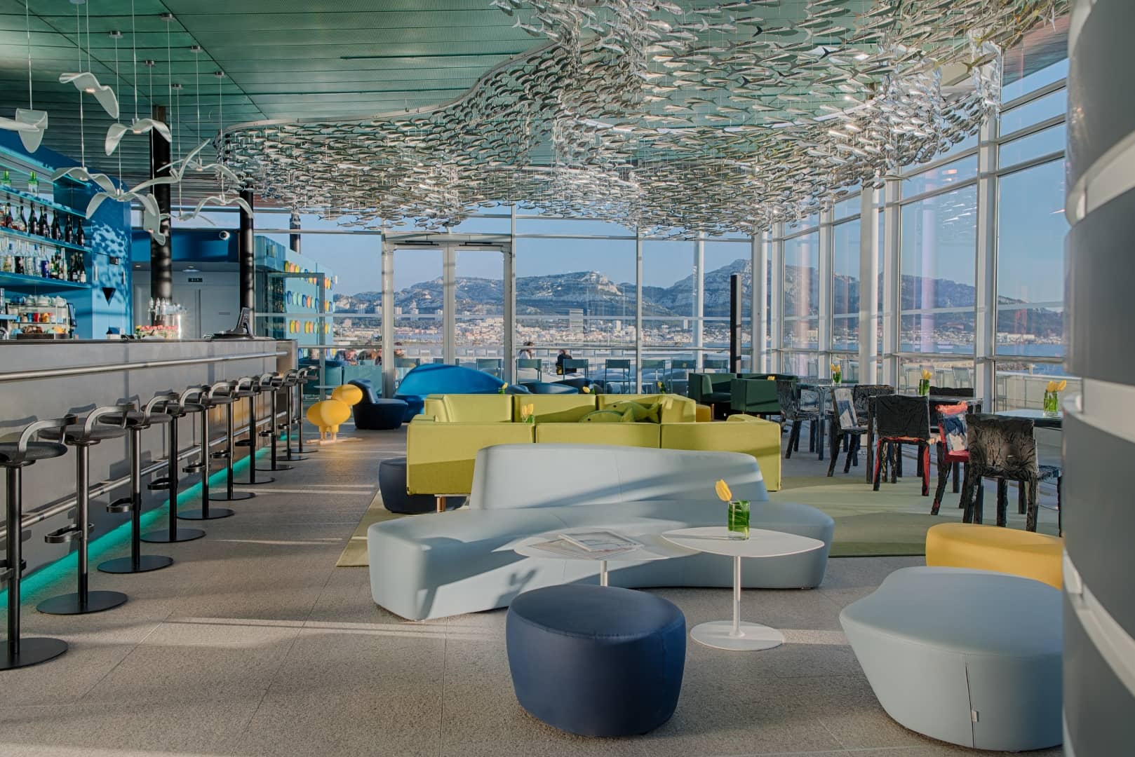 Lounge bar en rooftop propice à un moment afterwork, comprenant des canapés incurvés bleu pâle avec pouf bleu marine et petite table ronde au premier plan, une banquette modulable jaune-vert au centre, un comptoir de bar avec tabourets à gauche, et une nuée de sculptures de poissons suspendues au plafond avec vue sur mer et montagnes à travers de grandes baies vitrées en arrière-plan.