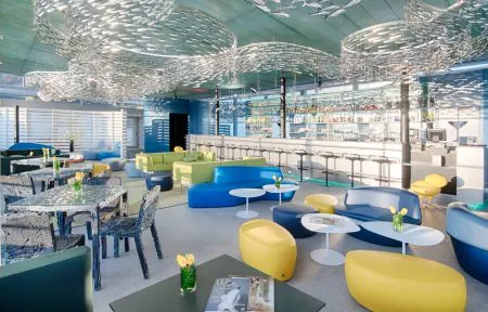 Salon d'hôtel pour afterwork en vue générale avec des canapés bleus, verts et jaunes autour de tables basses blanches au premier plan, un long bar avec des tabourets hauts à l'arrière-plan à droite, des tables de restauration transparentes avec des chaises à motif à gauche, et des installations décoratives suspendues argentées au plafond.