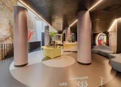 Hall d'hôtel plantant le décor d'une location de salle, comprenant une table ronde jaune décorée avec petite plante et objets au premier plan, de hautes colonnes arrondies roses et une signalétique au sol indiquant « auditorium » et « spa » au centre, et des canapés incurvés gris près d'une arche éclairée en rouge en arrière-plan, avec un mur en pierre naturelle visible à gauche.