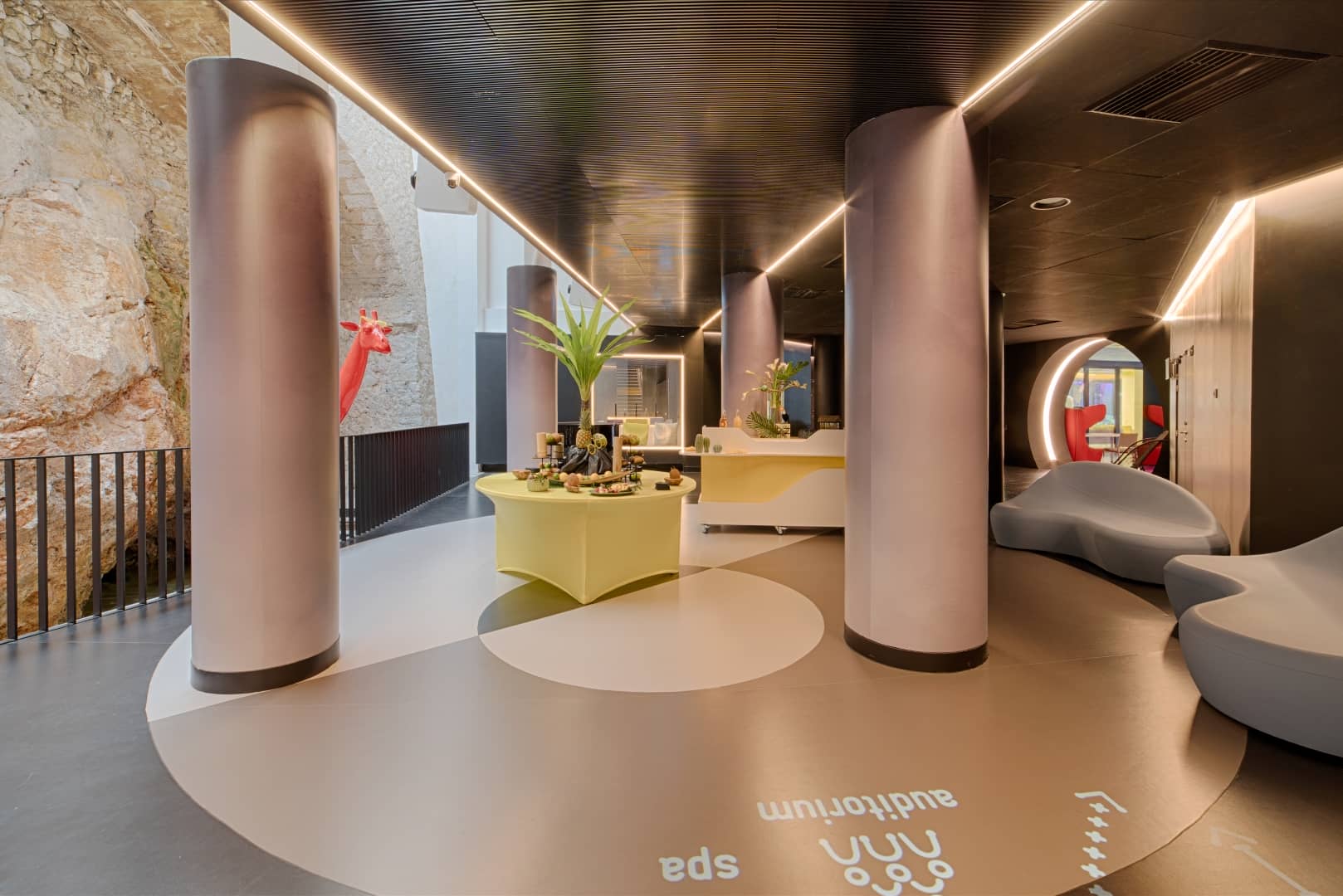 Hall d'hôtel plantant le décor d'une location de salle, comprenant une table ronde jaune décorée avec petite plante et objets au premier plan, de hautes colonnes arrondies roses et une signalétique au sol indiquant « auditorium » et « spa » au centre, et des canapés incurvés gris près d'une arche éclairée en rouge en arrière-plan, avec un mur en pierre naturelle visible à gauche.
