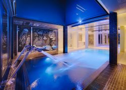 Bassin spa intérieur évoquant une ambiance séminaire bien-être, comprenant une rambarde chromée près d'un jet d'eau éclairé en bleu au premier plan, un mur en mosaïque de créatures marines en noir et blanc au centre, et un espace piscine lumineux avec marches carrelées visible à travers une arche en arrière-plan.